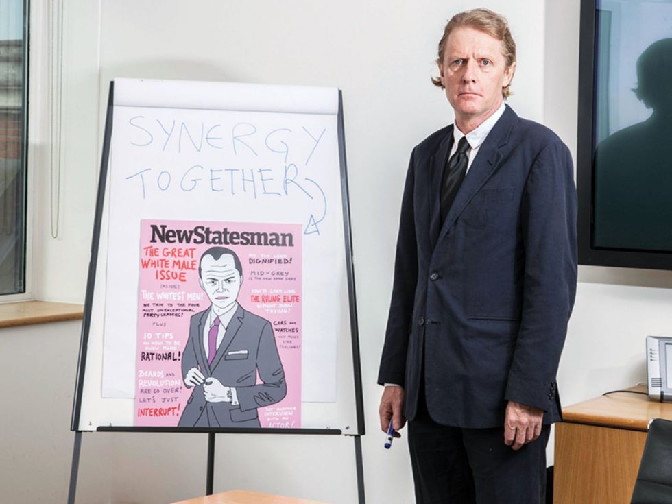 A light skinned man in a suit stands next to a white flip board with a copy of a New Statesmen cover for The Default Man article.