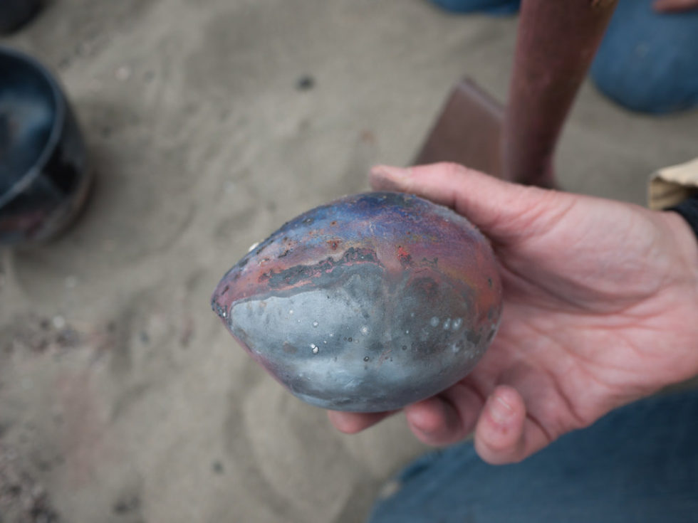 A hand over sand holding a ceramic egg streaked with black and gray and red